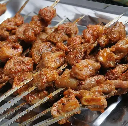 Uyghur cook grilling lamb shashlik skewers over open charcoal at a Xinjiang night market at dusk